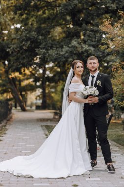 Bir gelin ve damat parkta fotoğraf için poz veriyorlar. Gelin beyaz bir elbise giyiyor ve damat siyah bir takım elbise giyiyor. Bir buket çiçek ve küçük bir buket çiçek tutuyorlar.
