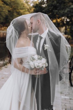 Bir gelin ve damat peçenin altında öpüşüyor. Gelin beyaz bir elbise giyiyor ve damat siyah bir takım giyiyor.