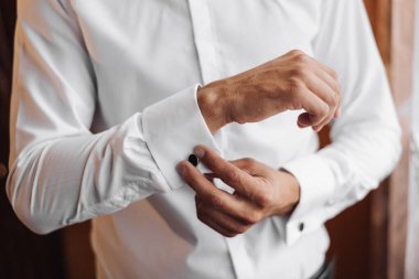 Bir adam beyaz gömlek ve siyah kol düğmesi giyiyor. Doğru yerde olduklarından emin olmak için gömleğini ve kol düğmelerini dikkatlice ayarlıyor. Detaylara dikkat etme kavramı