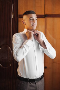 Bir adam beyaz gömlek ve siyah pantolon giymeye hazırlanıyor. Kravatını düzeltiyor ve kameraya bakıyor.