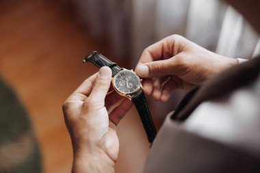 Bir adam bir saat tutuyor ve ona bakıyor. Saat siyah ve altındır. Adam takım elbise giyiyor.