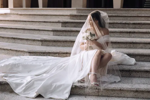 Gelinlik giymiş bir kadın merdivenlerde oturur. Elinde bir buket çiçek ve duvak tutuyor. Zarafet ve kutlama kavramı