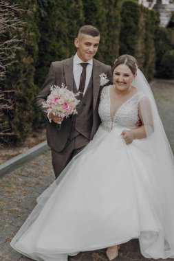 Bir gelin ve damat fotoğraf için poz veriyor. Gelin beyaz bir elbise giyiyor ve damat bir takım elbise giyiyor. Gelin elinde bir buket çiçek tutuyor.