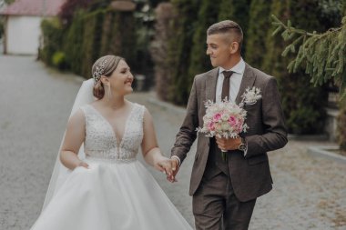 Bir gelin ve damat el ele tutuşup gülümsüyor. Gelin beyaz bir elbise giyiyor ve damat da takım elbise giyiyor.