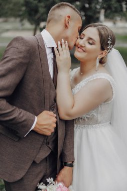 Bir gelin ve damat birbirlerinin alnını öpüyor. Gelin beyaz bir elbise giyiyor ve damat da takım elbise giyiyor.