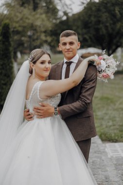 Bir gelin ve damat bir ağacın önünde fotoğraf için poz veriyorlar. Gelin beyaz bir elbise giyiyor ve damat da takım elbise giyiyor.