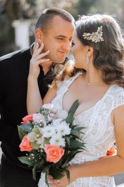 Bir erkek ve bir kadın bir buket çiçek tutarken birbirlerine sarılıyorlar. Adam siyah bir gömlek giyiyor ve kadın beyaz bir elbise giyiyor. Sahne romantik ve samimi.