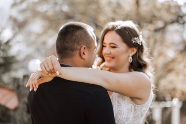 Bir erkek ve bir kadın birbirine sarılıyor, parmağında yüzük olan bir adamla. Kadın beyaz bir elbise giyiyor ve saçı örgülü. Sahne mutlu ve sevgi dolu.