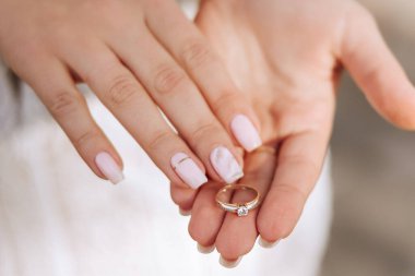 Bir kadın elinde bir yüzük tutuyor. Yüzük altından yapılmış ve üzerinde bir elmas var. Kadının elleri temiz ve bakımlı, ve beyaz bir elbise giyiyor.