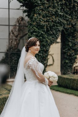 Beyaz elbiseli bir kadın yeşil bir çalılığın önünde duruyor. Elinde bir buket çiçek tutuyor.