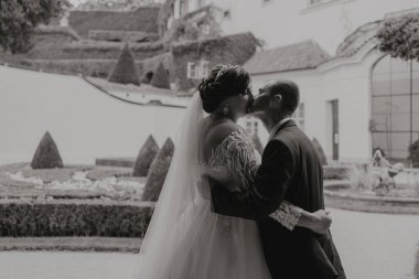 Gelin ve damat beyaz bir binanın önünde öpüşürler. Gelin peçe takıyor ve damat takım elbise giyiyor.