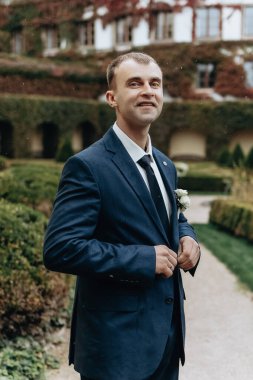 Mavi takım elbiseli ve beyaz gömlekli bir adam bir binanın önünde duruyor. O cebinde bir kravat ve beyaz bir çiçek taşıyor.