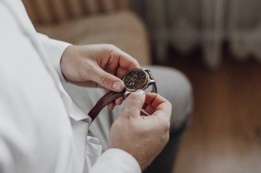 Bir adam saat takıyor ve ayarlıyor. Saat kahverengi ve gümüş bir şeridi var.