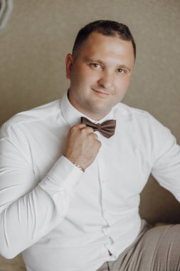 Beyaz gömlekli ve kahverengi papyonlu bir adam kanepede oturuyor. Gülümsüyor ve kameraya bakıyor.