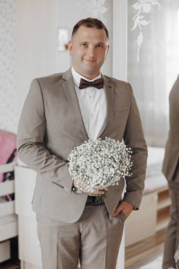 Takım elbiseli bir adam elinde bir buket beyaz çiçek tutuyor. Gülümsüyor ve kameraya bakıyor.