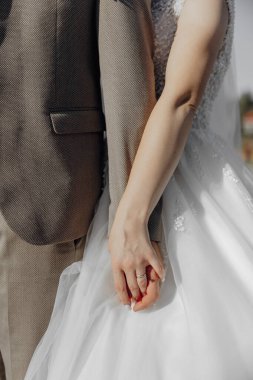 Bir adam ve bir kadın düğün fotoğrafında el ele tutuşuyorlar. Adam takım elbise giyiyor ve kadın beyaz bir elbise giyiyor. Fotoğraf, çift arasında bir sevgi ve bağlılık anı yakalıyor.