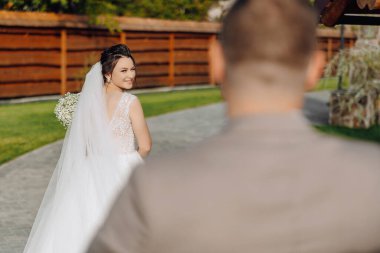 Bir gelin elinde bir buket çiçekle bir patikada yürüyor. Arkasında bir damat duruyor, ona bakıyor.