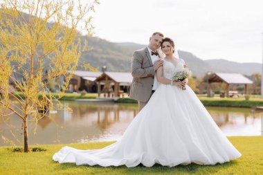 Bir gelin ve damat gölün önünde fotoğraf için poz veriyorlar. Gelin beyaz bir elbise giyiyor ve damat bir takım elbise giyiyor. Sahne huzurlu ve romantik, çift mutlu görünüyor.