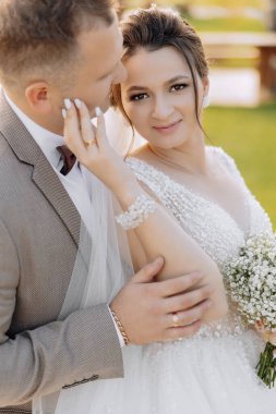 Bir gelin ve damat fotoğraf için poz veriyor. Gelin beyaz bir elbise giyiyor ve damat bir takım elbise giyiyor. İkisi de gülümsüyor ve kameraya bakıyor.