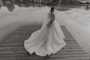 Rıhtımda elinde bir buket çiçekle bir gelin duruyor. Beyaz bir elbise ve duvak takıyor. Sahne huzurlu ve huzurlu.