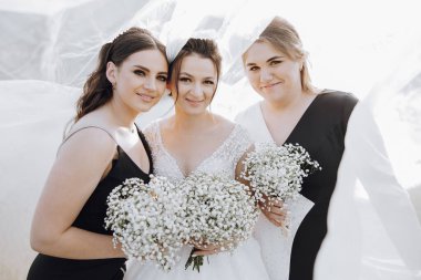 Üç kadın fotoğraf için poz veriyor, her biri bir buket beyaz çiçek tutuyor. Kadınlar siyah-beyaz giyinmiş, ve sahnenin klasik bir havası var.