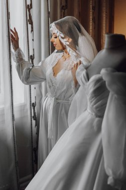 Beyaz elbiseli bir kadın pencerenin önünde duruyor. Duvak ve kolye takıyor.