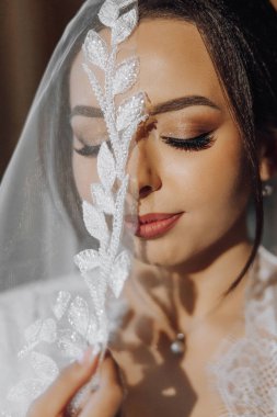 Bir kadın yaprak desenli bir peçe takıyor. Beyaz bir elbise giyiyor ve saçını arkaya çekiyor.