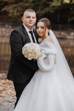 Bir gelin ve damat çitin önünde fotoğraf için poz veriyorlar. Gelin beyaz bir elbise giyiyor ve elinde bir buket çiçek tutuyor. Sahne mutlu ve kutlama dolu.