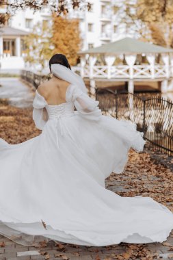 Beyaz elbiseli bir kadın yerde yaprakları olan bir yolda yürüyor. Elbise uzun ve dalgalı, ve kadın yürüyüşünün tadını çıkarıyor.