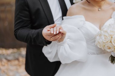 Gelin ve damat el ele tutuşuyor ve gelin beyaz bir elbise giyiyor.