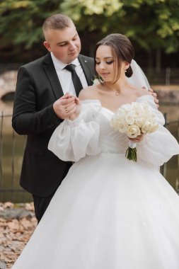 Bir gelin ve damat bir köprünün önünde fotoğraf için poz veriyorlar. Gelin elinde bir buket beyaz çiçek tutuyor.