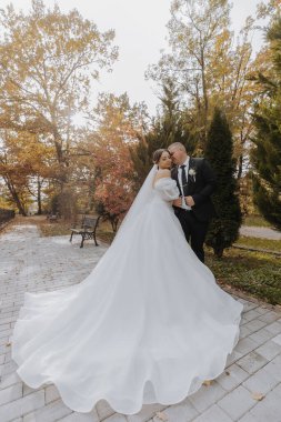 Bir gelin ve damat bir bankın önünde fotoğraf için poz veriyorlar. Gelin uzun beyaz bir elbise giyiyor ve damat bir takım elbise giyiyor. Sahne mutlu ve romantik.