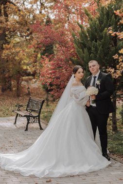 Bir gelin ve damat parkta bir bankın önünde duruyor. Gelin beyaz bir elbise giyiyor ve damat siyah bir takım giyiyor.