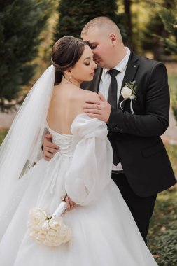 Bir gelin ve damat parkta birbirini kucaklıyor. Gelin beyaz bir elbise giyiyor ve damat siyah bir takım elbise giyiyor. Gelin elinde bir buket çiçek tutuyor.