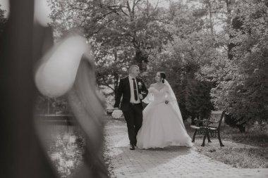 Bir gelin ve damat parkta bir patikada yürüyorlar. Gelin beyaz bir elbise giyiyor ve damat bir takım elbise giyiyor. El ele tutuşup kameraya gülümsüyorlar. Sahne mutlu ve romantik.