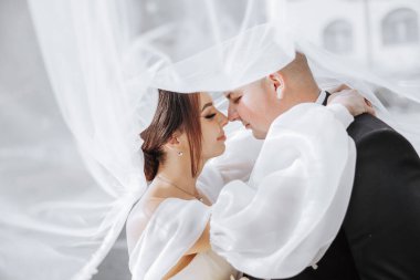 Bir gelin ve damat beyaz bir peçenin altında birbirlerini kucaklıyorlar. Gelin kolye takıyor ve damat kravat takıyor. Sahne romantik ve samimi.