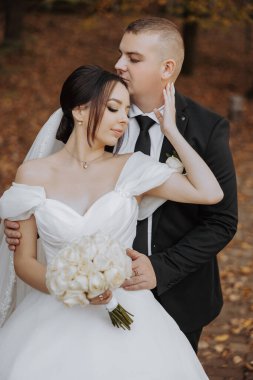 Bir gelin ve damat ormanda birbirini kucaklıyor. Gelin beyaz bir elbise giyiyor ve damat siyah bir takım elbise giyiyor. Gelin elinde bir buket çiçek tutuyor.