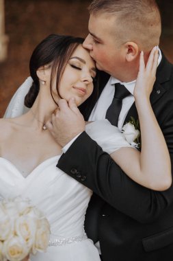 Gelin ve damat birbirlerini kucaklıyor, gelin beyaz elbise giyiyor ve damat siyah takım elbise giyiyor.