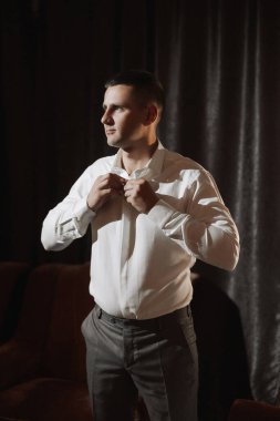 Beyaz gömlekli bir adam giyiniyor. O bir perdenin önünde duruyor. Adam gri takım elbise ve gri pantolon giyiyor.