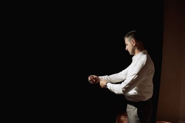 Beyaz gömlekli bir adam karanlık bir odada duruyor, saatine bakıyor. Acelesi var ve sık sık saati kontrol ediyor.