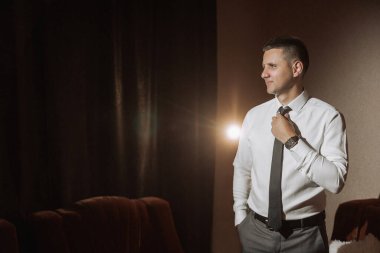 Takım elbiseli ve kravatlı bir adam perdenin önünde duruyor. Kravatını düzeltiyor ve kameraya bakıyor. Formalite ve profesyonellik kavramı. Çünkü adam takım elbise ve kravat giymiş.