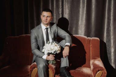 Takım elbiseli bir adam elinde bir buket çiçekle kanepede oturur. Zarafet ve incelik kavramı, adam resmi bir takım elbise giymiş ve elinde güzel bir buket çiçek tutuyor.