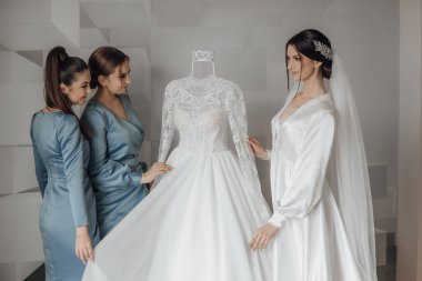 Beyaz bir gelinliğin önünde üç kadın duruyor. Kadınlardan biri elbiseyi diğer ikisine doğru tutuyor. Sahne, yaklaşan düğün için heyecan ve beklentilerden biridir.
