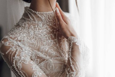 Bir kadın dantel yakalı ve inci kolyeli beyaz bir elbise giyiyor. Eli boynunda kameraya poz veriyor. Elbise uzun ve zarif.