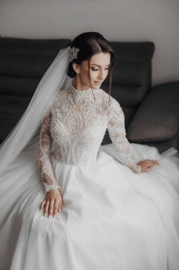 Beyaz gelinlikli bir kadın kanepede oturur. Duvak ve beyaz dantelli bir elbise giyiyor.