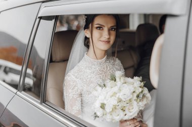 Bir gelin elinde bir buket beyaz çiçekle arabada oturuyor. Gülümsüyor ve pencereden dışarı bakıyor.