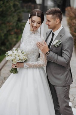 Bir gelin ve damat bir buket beyaz çiçekli bir resim için poz veriyorlar. Gelin beyaz bir elbise giyiyor ve damat gri bir takım elbise giyiyor. Sahne mutlu ve kutlama dolu.