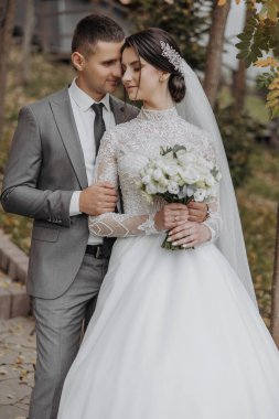 Gelin ve damat birlikte duruyorlar, gelin elinde bir buket çiçek tutuyor. Gelin beyaz bir elbise giyiyor ve damat bir takım elbise giyiyor. Sahne mutlu ve romantik.