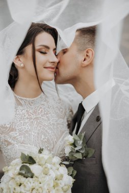 Bir gelin ve damat beyaz bir peçenin altında öpüşüyorlar. Gelin beyaz bir elbise giyiyor ve damat bir takım elbise giyiyor. Sahne samimi ve romantik, aşkı yakalıyor.