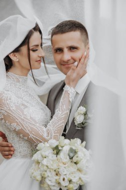 Gelin ve damat, gelinin kafasında beyaz bir peçeyle birbirlerini kucaklıyorlar. Gelin bir buket çiçek tutuyor ve damat elini tutuyor. Sahne bir düğün fotoğrafı.